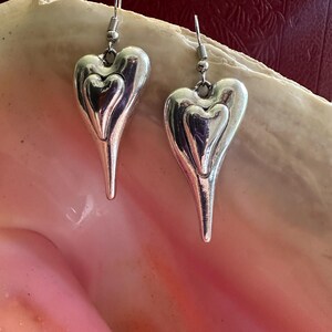May include: A pair of silver heart-shaped earrings. Each earring features a larger heart with a smaller heart nested inside. The earrings have a long, pointed bottom and are suspended from a silver hook.