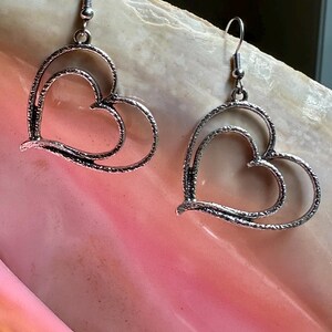 May include: A pair of silver-toned heart-shaped earrings. Each earring features a double heart design with a textured finish. The earrings hang from a simple hook.