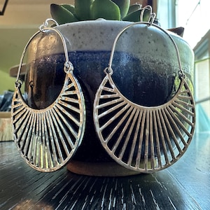 May include: Silver-toned, fan-shaped earrings with a linear design. The earrings have a half-circle shape with a series of vertical lines radiating from the center. The earrings are displayed on a dark wooden surface.