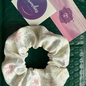 May include: A white fabric hair scrunchie featuring a repeating pattern of snowflakes in shades of pink, blue, and gold. Included are two branded cards with the text Scrunchies by Chelsea, one circular and purple, the other rectangular and pink.