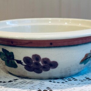May include: A ceramic bowl with a white interior and a cream-colored rim. The exterior features a band of brown and a design of green leaves and purple grapes. The bowl is likely for serving food.