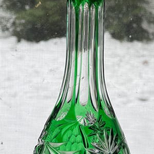 May include: A green glass decanter with a faceted crystal stopper. The decanter features intricate cut glass designs of leaves, grapes, and starburst patterns. The clear glass stopper adds a touch of elegance to this decorative piece.