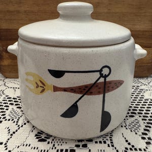 May include: White ceramic canister with a speckled finish and a lid. The canister has a brown, black, and yellow design of kitchen utensils.