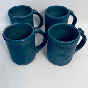 May include: A set of four matching mugs in a deep, matte blue finish. Each mug features a cylindrical body, a rounded rim, and a curved handle with a small, raised thumb rest detail at the top. The surface has a subtle, textured appearance.