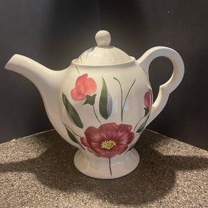May include: White ceramic teapot with a floral design. The teapot has a round body with a spout and a handle. The floral design features pink, red, and green flowers.