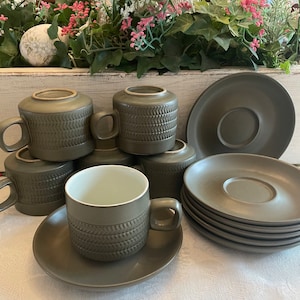 May include: A set of olive green ceramic teacups and saucers. The cups have a textured, woven pattern around the middle and a handle. The saucers are round and have a recessed center. The set is arranged on a white tablecloth.