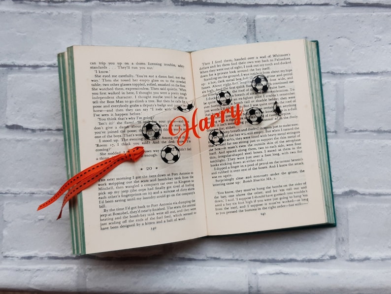 May include: A personalized bookmark with the name "Harry" printed in orange on a clear plastic background. The bookmark features a design of soccer balls and a red ribbon.