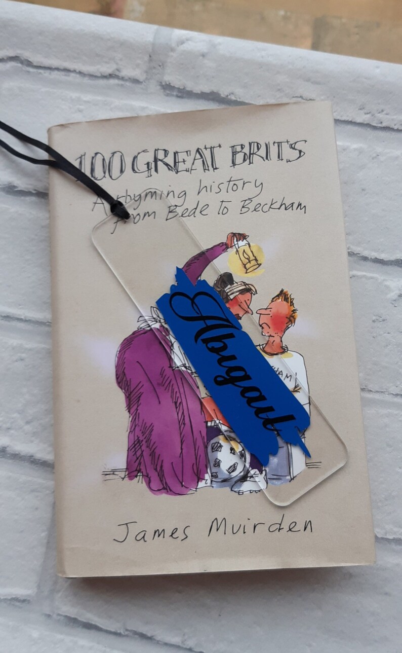 May include: A blue and white bookmark with the word "August" written in black cursive. The bookmark is on top of a book titled "100 Great Brits A rhyming history from Bede to Beckham" by James Muirden.
