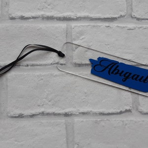 May include: A clear acrylic bookmark with a blue paint-splatter design and the name "Abigail" written in black cursive.