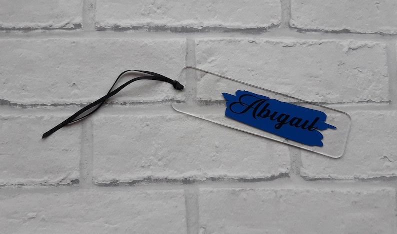 May include: A clear acrylic bookmark with a blue paint-splatter design and the name "Abigail" written in black cursive.