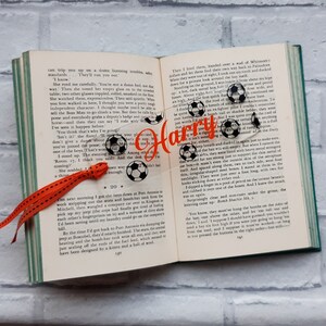 May include: A clear acrylic bookmark with a black and white soccer ball design and the name "Harry" in orange text. The bookmark has a red ribbon attached to it.