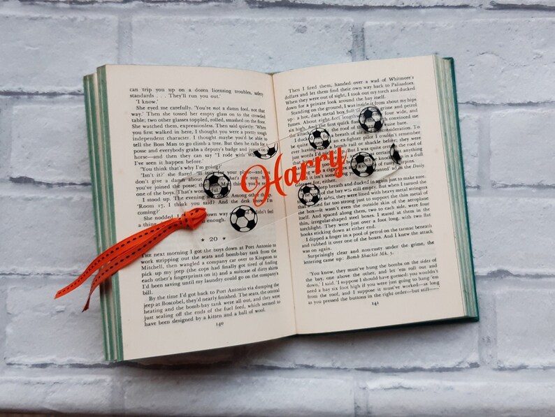 May include: A clear acrylic bookmark with a black and white soccer ball design and the name "Harry" in orange text. The bookmark has a red ribbon attached to it.