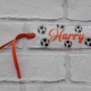 May include: A clear acrylic bookmark with a black and white soccer ball pattern and the name "Harry" in orange lettering. The bookmark has a bright orange ribbon attached.