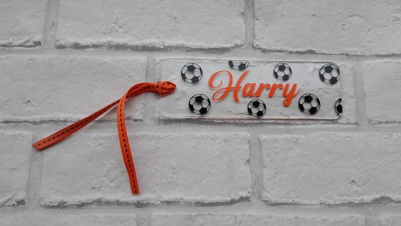 May include: A clear acrylic bookmark with a black and white soccer ball pattern and the name "Harry" in orange lettering. The bookmark has a bright orange ribbon attached.