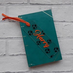 May include: A clear acrylic bookmark with a soccer ball design and the name "Harry" in orange lettering. The bookmark is attached to a teal book with a ribbon.