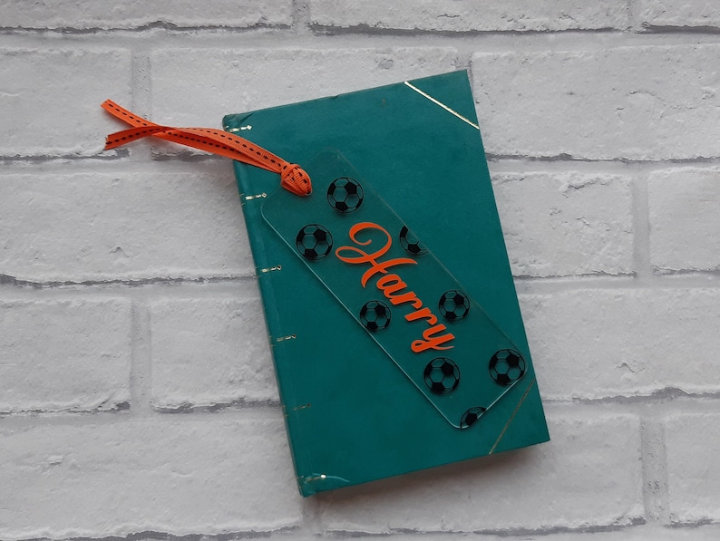 May include: A clear acrylic bookmark with a soccer ball design and the name "Harry" in orange lettering. The bookmark is attached to a teal book with a ribbon.
