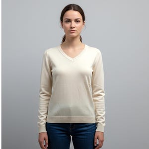 Può includere: Un maglione color crema con scollo a V e maniche lunghe. Il maglione è abbinato a jeans blu. La modella è centrata davanti a uno sfondo grigio chiaro. Il maglione ha orlo e polsini a coste.