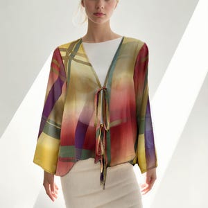 May include: A colorful, sheer kimono-style top with a tie closure. The top features a vibrant plaid pattern in shades of yellow, red, purple, and green. The model is wearing a white top and a cream-colored skirt.