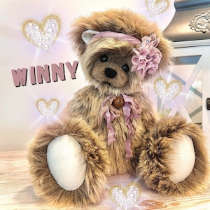 Puede incluir: Un oso de peluche marrón con una cinta y una flor rosas. El oso tiene una campana alrededor del cuello y la palabra "WINNY" se muestra en rosa. Corazones decorativos con detalles de perlas están dispersos alrededor del oso.