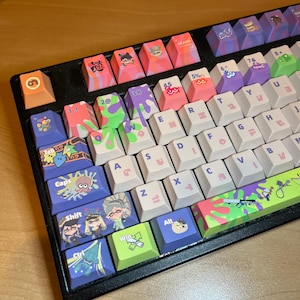 May include: A black mechanical keyboard featuring custom keycaps with colorful Splatoon-themed graphics, including character illustrations, paint splatters, and icons. The keys display standard QWERTY letters and symbols in a light pink font.
