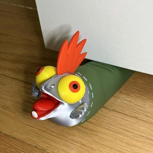 May include: A doorstop shaped like a stylized fish with a green body, a silver head, large yellow eyes with black pupils, a bright orange fin on top, and an open mouth with a red tongue and a single white tooth protruding from the lower jaw.