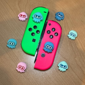 May include: A neon green and a neon pink video game controller, each featuring a small squid-shaped joystick cover. Six additional squid-shaped joystick covers in light blue and light pink are arranged around the controllers on a wooden surface.