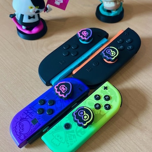 May include: Four Nintendo Switch Joy-Cons in black, blue, green, and orange. Each controller has a unique color scheme and features a matching thumbstick cover with a cartoon squid design. Two small figurines of cat-like characters are also present.