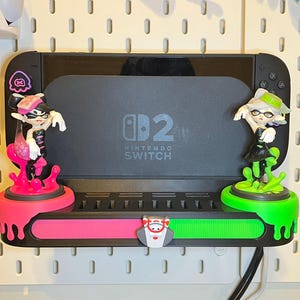 May include: A black Nintendo Switch console with a custom dock featuring two character figurines. The dock has a pink and green colour scheme with a small character in the centre. The figurines are of characters from the game.