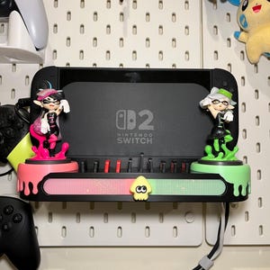 May include: A black Nintendo Switch console with two Inkling figures on a custom shelf. The shelf is black, pink, and green with a glittery section and a squid icon. The console is mounted on a white pegboard.