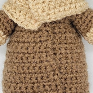 May include: A crocheted brown and beige doll wearing a brown hooded robe. The doll has a green head and is crocheted in a detailed stitch.