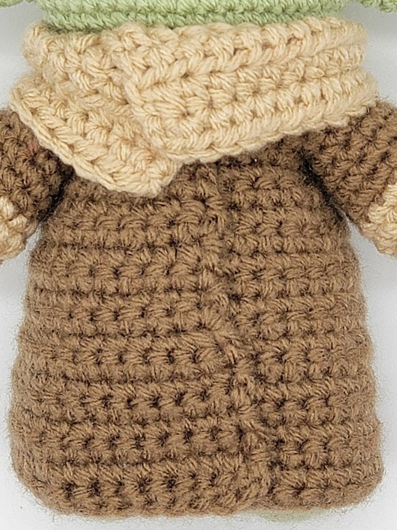 May include: A crocheted brown and beige doll wearing a brown hooded robe. The doll has a green head and is crocheted in a detailed stitch.