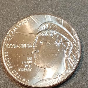 May include: A silver coin featuring a profile of a person with flowing hair and a radiant design. The coin is inscribed with "UNITED STATES OF AMERICA", "1776-2026", and "IN GOD WE TRUST". The coin has a detailed edge and a reflective surface.