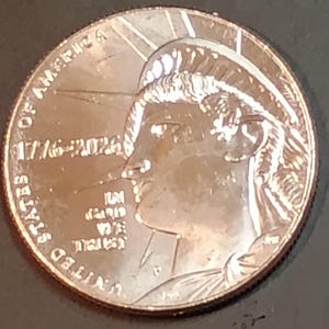 May include: A silver coin featuring a profile of a person with a crown and the text "UNITED STATES OF AMERICA" and "1776-2026". The coin has a detailed design and a shiny finish. The coin is a commemorative piece.