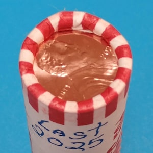 May include: A roll of pennies with a red and white striped paper wrapper. The wrapper has the text "EAST 50 25" written in blue ink. The pennies are visible at the top of the roll.