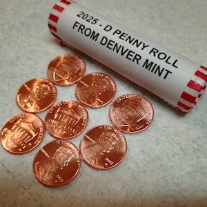 May include: A roll of 2025-D penny coins from the Denver Mint, with several loose pennies scattered around. The pennies are copper-colored, and the roll is white with red stripes on the ends and black text.