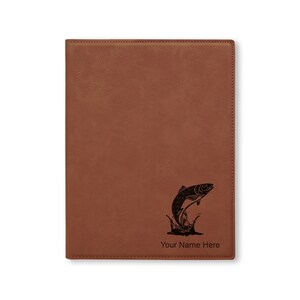 May include: Brown leather journal with a black engraved image of a fish jumping out of the water. The journal has the text "Your Name Here" engraved below the image.