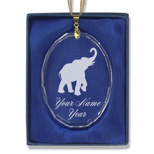 May include: A clear glass oval ornament with a white silhouette of an elephant. The ornament has a gold chain and is in a blue velvet box. The ornament is personalized with "Your Name Year".