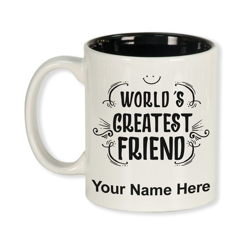K&ouml;nnte beinhalten: Wei&szlig;er Keramikbecher mit schwarzem Innenraum und gebogenem Henkel. Der Becher zeigt den Text "WORLD'S GREATEST FRIEND" in dekorativer schwarzer Schrift, mit Platz f&uuml;r einen personalisierten Namen darunter.