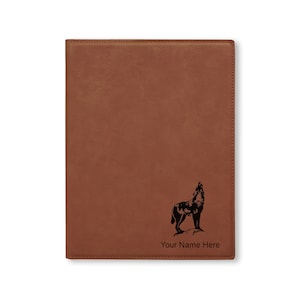 May include: A brown leather journal with a howling wolf graphic and the text "Your Name Here". The journal has a stitched border and a smooth, textured surface. The wolf is black.