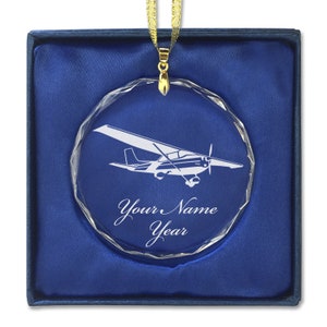 May include: A clear crystal ornament with a white silhouette of a small airplane. The ornament is in a blue velvet box and has a gold ribbon attached. The ornament is personalized with the text "Your Name Year".