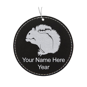 May include: A round, black ornament with a silver squirrel design. The ornament has white stitching around the edge and a silver string for hanging. The text "Your Name Here" and "Year" are printed in white.