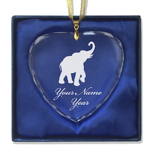 May include: A white elephant silhouette is etched into a clear glass heart-shaped ornament. The ornament is in a blue velvet box with a gold ribbon. The text "Your Name Year" is engraved below the elephant.