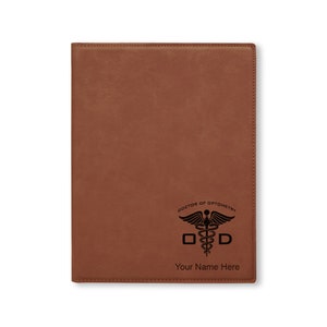 May include: Brown leather notebook with a black engraved design of a caduceus, the symbol of medicine, with the text "Doctor of Optometry" and "ODD" above the caduceus and "Your Name Here" below.