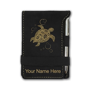 May include: Black leatherette notebook with a gold-colored sea turtle design and the text "Your Name Here". The notebook has a black elastic band closure and a silver pen.