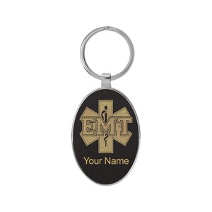 May include: A black oval keychain with a gold star of life and the letters EMT. The keychain has a silver ring and the text "Your Name" is printed below the star of life.