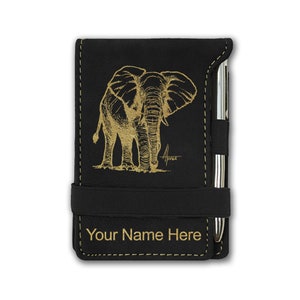 May include: Black notepad with a gold elephant illustration and the text "Your Name Here". The notepad has a pen holder and an elastic band closure. The elephant design is detailed and artistic.