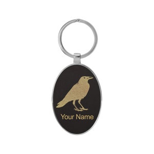 May include: A black oval keychain with a gold silhouette of a crow and the text "Your Name".
