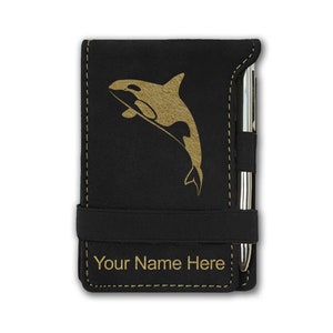 May include: Black notepad with a gold orca design and the text "Your Name Here". The notepad has a pen holder and an elastic band closure. The stitching is gold.