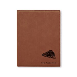 May include: A brown leather journal with a black silhouette of a train and the text "Your Name Here" engraved on the front.