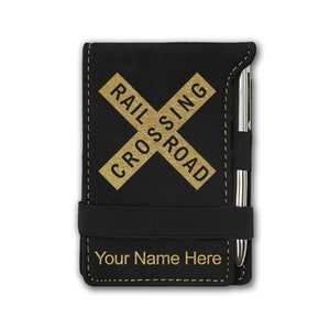 May include: Black leather notebook with a gold railroad crossing sign and the text "Your Name Here". The notebook has a pen holder and a black elastic band.